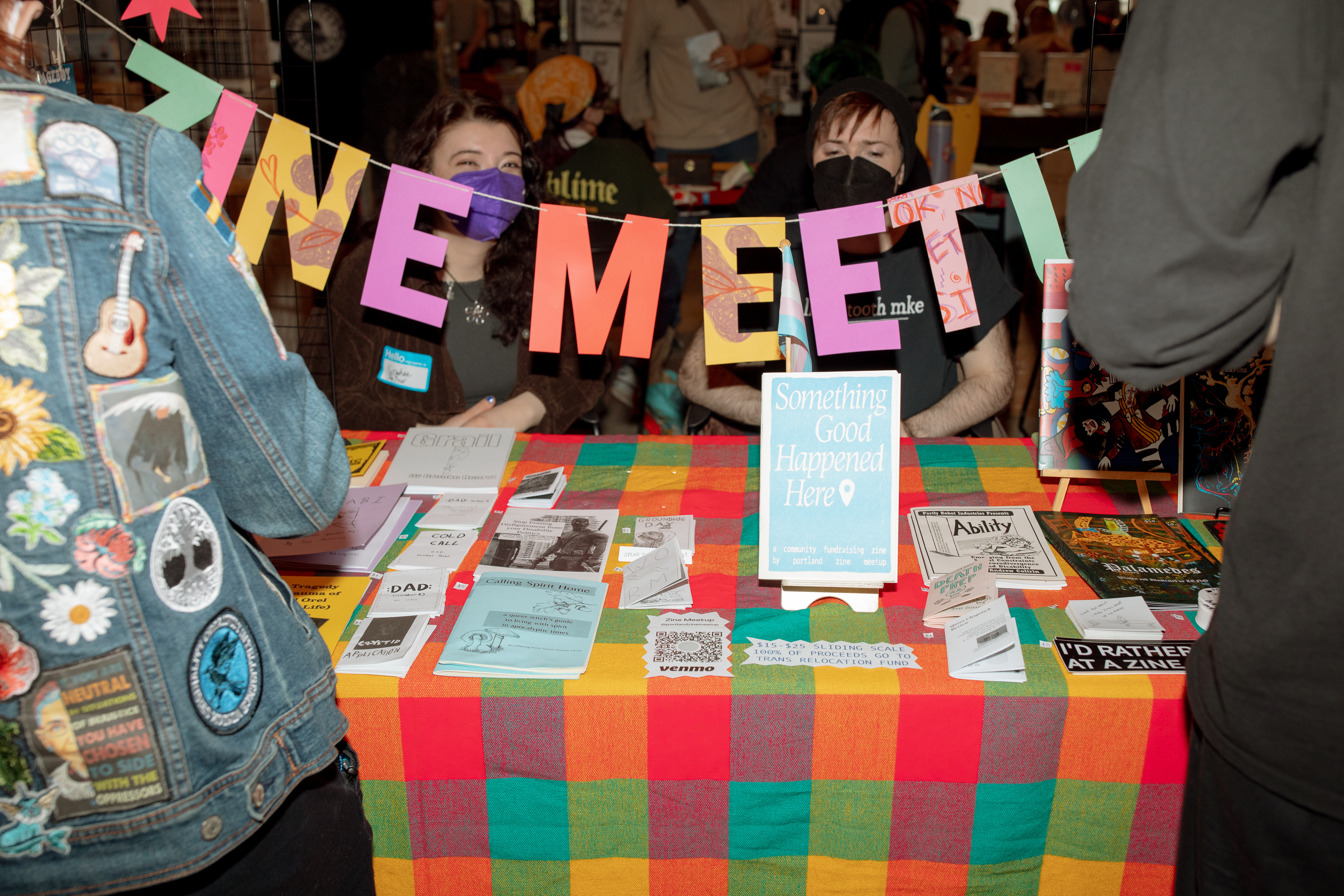 image of two people sitting behind a banner that says Zine Meetup in front of a table of zines