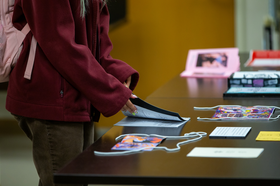 The hands and trunk of a person in a maroon sweatshirt with colorful fingernails opens a zine, with other zines on the table. The title and content of the zines are blurry and not visible.