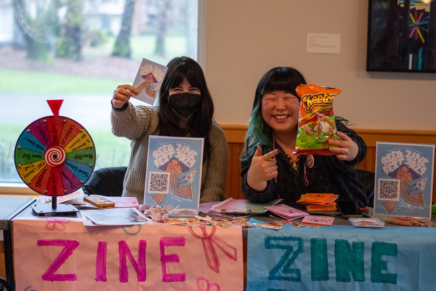 Two people, smiling, one in a mask, sit at a table with a colorful spinning wheel, stickers, candy, and zines. One person holds a paper that reads "Reed Zine Fest 2024" in cloud-like font, and the other holds and points to a bag of cheetos.