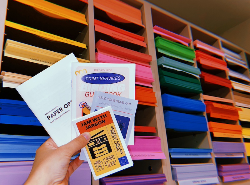 Brightly colored paper on shelving in the background, wuth a hand holding four zines titled: "Jam with Jargon: understanding print lingo", "Bleed your heart out", "Reed College Print Services Binding", and "Introduction to paper"