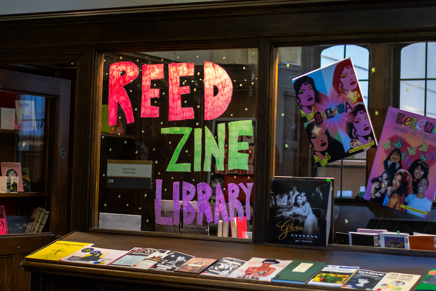 Dark wood shelving holds zines, whose titles and covers are blurry. The words Reed Zine Library are written in red, green, and purple free hand letting on a window, along with gold stars. Several images or record covers of Selena are attached to the window on the right.